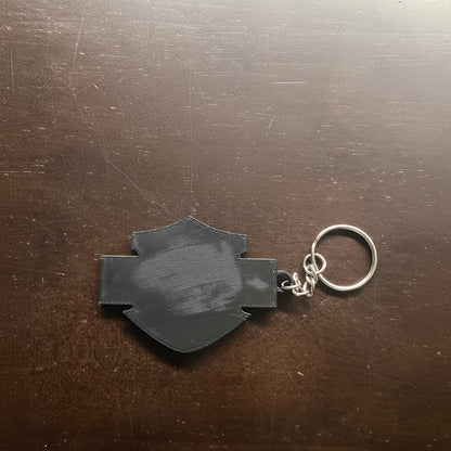 Black 3D-printed keychain on a wooden surface with 'Whatcha Printing' logo in the background.