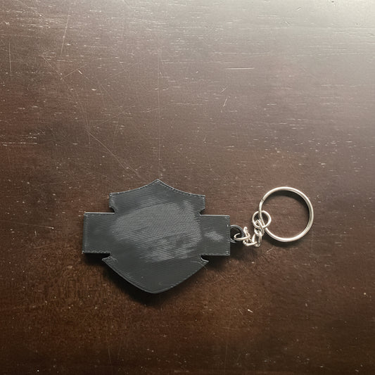 Black 3D-printed keychain on a wooden surface with 'Whatcha Printing' logo in the background.